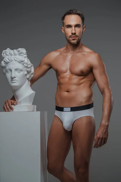A stunning male model with a chiseled torso and in underwear striking a pose next to a bust of an ancient Greek sculpture