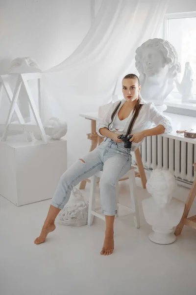 漂亮的雕塑家 身穿白衬衫和牛仔裤 头戴吊袜带 坐在雕塑家作坊的椅子上 手持相机 — 图库照片