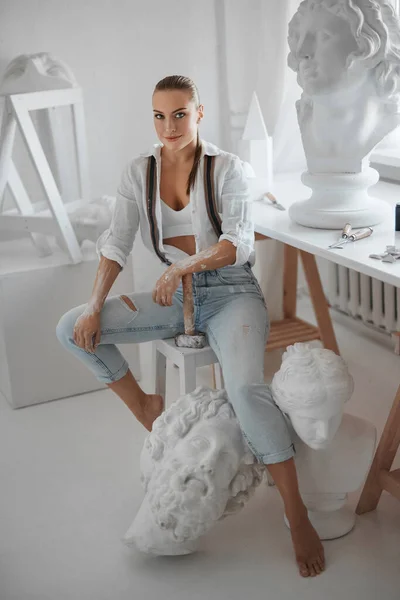 有才华的女雕塑家 身穿白衬衫和牛仔裤 头戴吊袜带 手持锤子 坐在雕塑家工作室的椅子上 四周都是雕塑品 — 图库照片