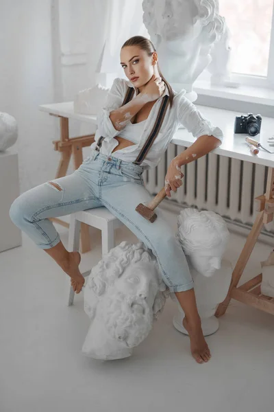 一位漂亮的女雕塑家 身穿宽松的白衬衫和牛仔裤 头戴吊带衫 坐在雕塑家工作室的凳子上 — 图库照片