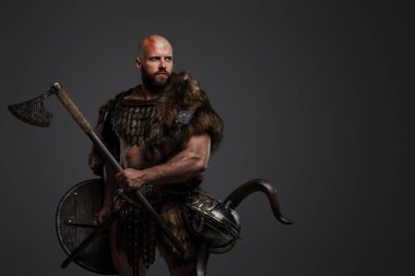 Ürkütücü, kel, sakallı, kürk ve hafif zırh giyen, kemerine bir kask takan, tehdit edici bir baltayı ve kalkanı gri bir arka planda tutan bir Viking.