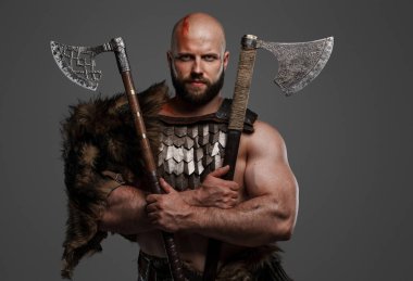 Kel, kaba, sakallı, kürk ve hafif zırh giymiş, kemerine bir kask takmış gri bir arka planda iki balta tutan bir Viking.