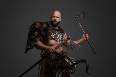 Kel ve sakallı, hayvan kürkü ve hafif zırh giymiş kemerinden sarkan bir miğferle gri bir arka plana kocaman bir balta sallayan güçlü ve korkutucu bir Viking.
