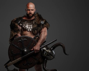 Tüylü, kel, sakallı, tüylü ve hafif zırhlı bir Viking, kemerine bir kask takılmış, gri arka planda bir balta ve kalkan tutuyor.