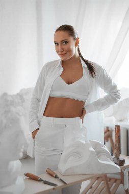 Güzel heykeltraş beyaz gömlek ve beyaz iç çamaşırlarıyla poz veriyor. Heykeltraş atölyesindeki antik Yunan heykelleriyle çevrili.