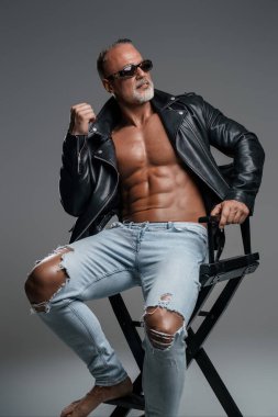 Şık gri sakallı, çıplak gövdeli, siyah deri ceketli, yırtık kot pantolonlu, stüdyo sandalyesinde otururken güneş gözlüklü, olgun, kaslı bir erkek model.