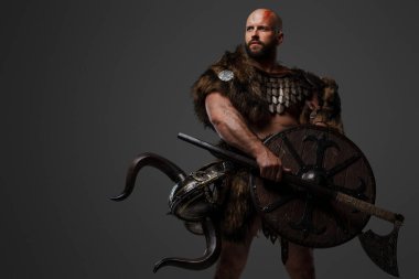 Tüylü, kel, sakallı, tüylü ve hafif zırhlı bir Viking, kemerine bir kask takılmış, gri arka planda bir balta ve kalkan tutuyor.