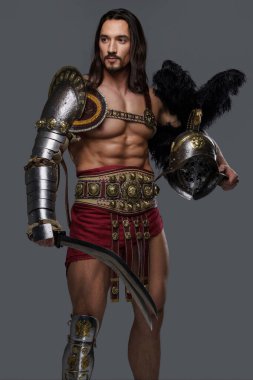 Muscular gladiator in graceful lightweight armor strikes a pose while holding a gladius and helmet, on a grey background clipart