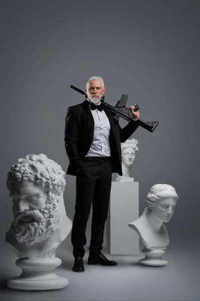 Stylish older man stands in a relaxed confident pose on a grey background, holding a gun, surrounded by three ancient sculptures, dressed in an expensive suit