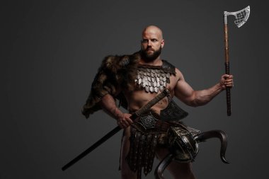 Sert, kaslı, sakallı, kürk ve hafif zırh giymiş, beline bir kask takmış gri bir arka plana karşı iki balta sallayan bir Viking savaşçısı.