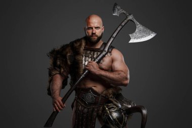 A strong and intimidating Viking man in a beard and bald head, dressed in animal fur and light armor, with a helmet hanging from his belt, brandishing a huge axe against a gray background clipart