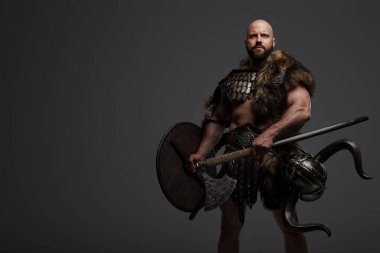 Tüylü, kel, sakallı, tüylü ve hafif zırhlı bir Viking, kemerine bir kask takılmış, gri arka planda bir balta ve kalkan tutuyor.