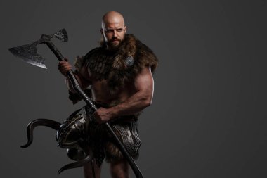 Kel ve sakallı, hayvan kürkü ve hafif zırh giymiş kemerinden sarkan bir miğferle gri bir arka plana kocaman bir balta sallayan güçlü ve korkutucu bir Viking.