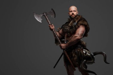 Kel ve sakallı, hayvan kürkü ve hafif zırh giymiş kemerinden sarkan bir miğferle gri bir arka plana kocaman bir balta sallayan güçlü ve korkutucu bir Viking.