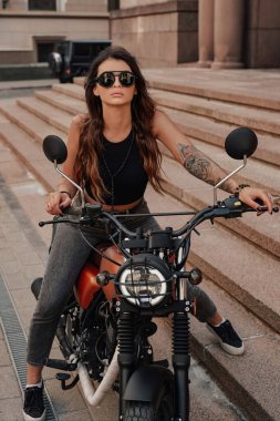 Kot pantolon ve kolunda güneş gözlüğü ve dövmesi olan şık bir esmer Avrupa 'da eski bir kaldırım taşı caddesindeki eski motosikletinde poz veriyor.