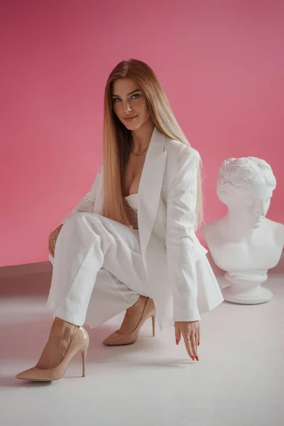 身着白色西服和性感胸罩的优雅模特 在一家粉红工作室里 穿着华丽的摄影装束 站在古希腊半身像旁边 — 图库照片