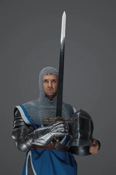 骄傲的骑士站在那里 拿着他的剑和头盔 穿着蓝色的外套 戴着链甲 身穿中世纪盔甲 背景是灰色的 — 图库照片