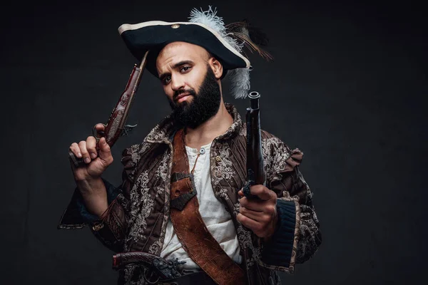 有魅力的海盗 留着黑色胡子 身穿背心 头戴帽子 拿着两支步枪 靠在黑暗的纹理墙壁上 — 图库照片