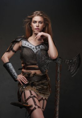 Göz kamaştırıcı Viking esinlenici kadın, iki eliyle baltayla dokulu gri bir duvara karşı poz veriyor, zincirli bir üst ve kürk etek giyiyor.