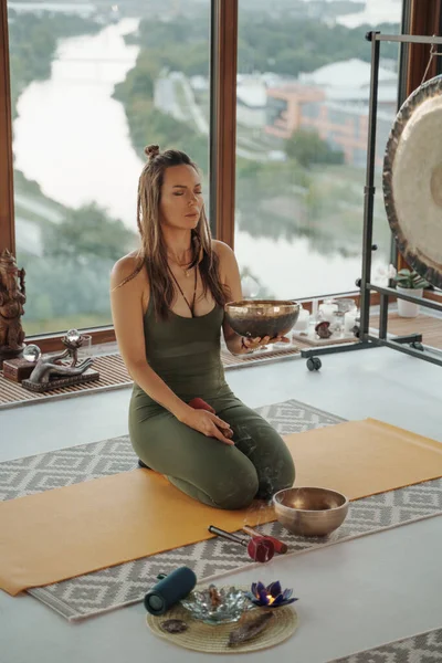Woman in sportswear practicing spiritual activities in a peaceful environment, meditating with a singing Tibetan bowl in an apartment overlooking a panoramic city skyline