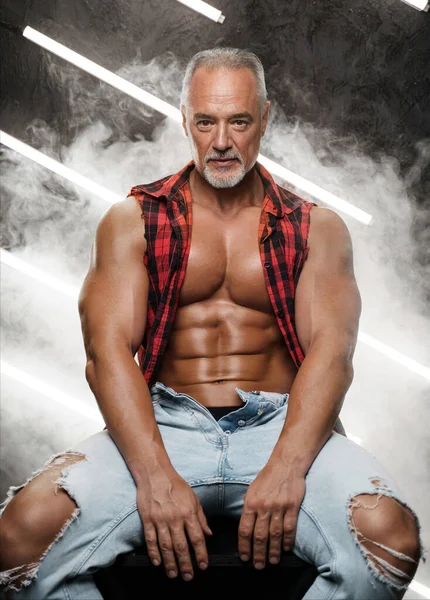 Masculine middle-aged male model with chiseled torso grey beard and bare chest, clad in an open plaid shirt and ripped jeans, seated on black cube surrounded by lights and smoke against textured wall