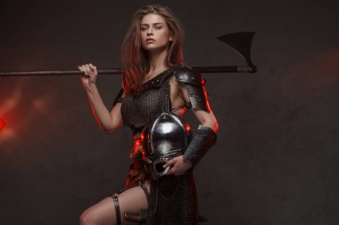 Göz kamaştırıcı Viking kadın. Zincirli bir üst ve kürk giymiş. Kırmızı ışıkla aydınlatılmış iki elli bir balta.