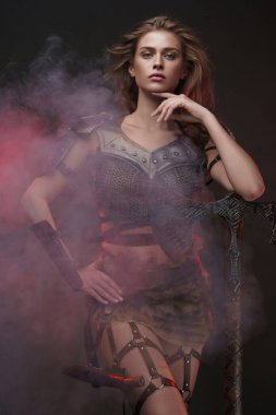 Zincirli tişört ve kürk etek giymiş muhteşem bir Viking kızı iki elli bir baltayla poz veriyor. Kırmızı ışık desenli gri bir duvara vuruyor.