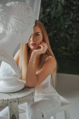Beyaz pantolon ve seksi bir sütyen giymiş şık ve hayat dolu bir kadın, heykeltraş atölyesinde antik bir Yunan büstünün yanında oturmuş, yaratıcı ve kentsel bir görünüm sergiliyor.