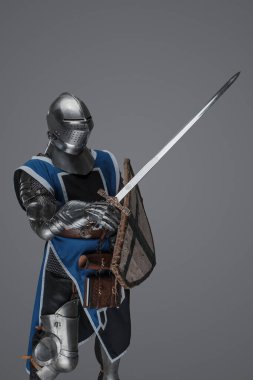 Brave medieval warrior dressed in armor and blue surcoat waving his sword energetically in battle, against a gray backdrop