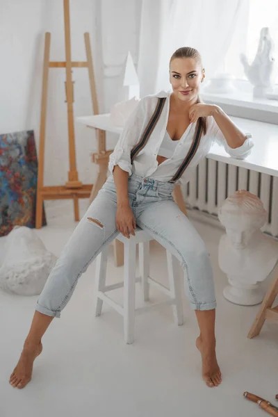 一位漂亮的女雕塑家 身穿宽松的白衬衫和牛仔裤 头戴吊带衫 坐在雕塑家工作室的凳子上 — 图库照片