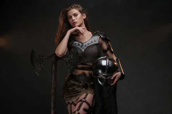 強力な斧とヘルメットでポーズする美しいヴァイキングの戦士モデルは テクスチャーされた背景に対する中世の印象的な衣装で強さと女性らしさを示しています — ストック写真