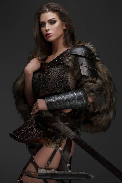 An attractive Viking-themed model in chainmail armor and fur, holding an axe against gray backdrop