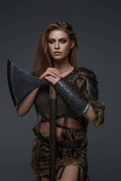 An attractive Viking-themed model in chainmail armor and fur, holding an axe against gray backdrop
