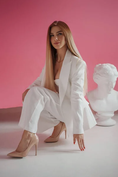 身着白色西服和性感胸罩的优雅模特 在一家粉红工作室里 穿着华丽的摄影装束 站在古希腊半身像旁边 — 图库照片