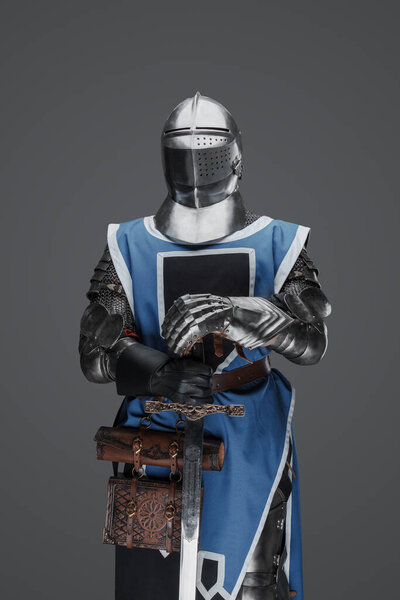 Средневековый солдат в синем сюртуке и доспехах, держащий меч в неподвижной позе, похожей на статую, на сером фоне
