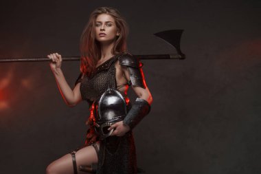 Göz kamaştırıcı Viking kadın. Zincirli bir üst ve kürk giymiş. Kırmızı ışıkla aydınlatılmış iki elli bir balta.