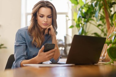 Çiçeklerle dolu bir odada dizüstü bilgisayarıyla çalışan, mavi gömlekli, gülümseyen serbest bir kadın.