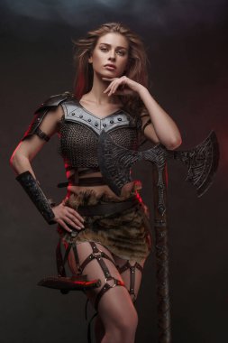 Zincirli tişört ve kürk etek giymiş muhteşem bir Viking kızı iki elli bir baltayla poz veriyor. Kırmızı ışık desenli gri bir duvara vuruyor.