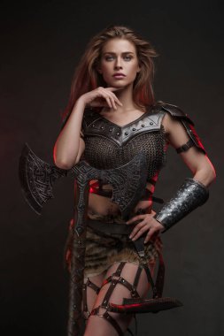 Zincirli tişört ve kürklü etek giyen büyüleyici Viking kız, çift elli baltayla dokulu gri bir duvara poz veriyor.