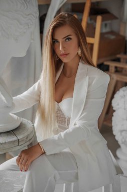 Modelimsi görünümlü çekici bir genç kadın, şık beyaz bir takım elbise ve tahrik edici bir sütyen giyiyor, antik bir Yunan büstünün yanındaki heykeltıraşlık atölyesinde otururken poz veriyor.