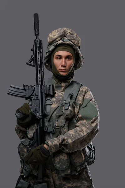 ユニフォームとヘルメットの若い兵士は 明白な灰色の背景に深刻な表現をポーズし 軍人の強さと献身を示しています — ストック写真