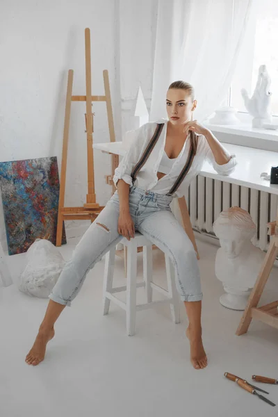 漂亮的雕塑家穿着一件白色的 没有扣子的衬衫和一条有吊带的牛仔裤 坐在她车间的椅子上 — 图库照片