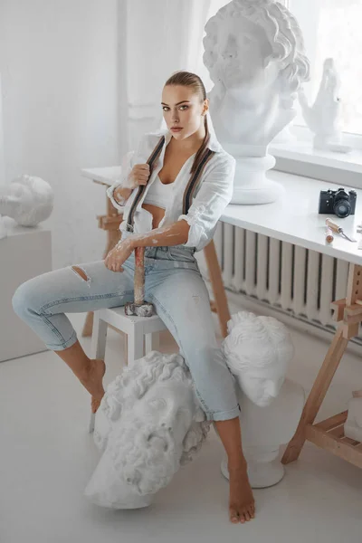 白いボタンなしのシャツとジーンズをサスペンサーで着た才能ある女性彫刻家は ハンマーを持ち 彫刻に囲まれた彼女の彫刻工房で椅子にポーズをかける — ストック写真