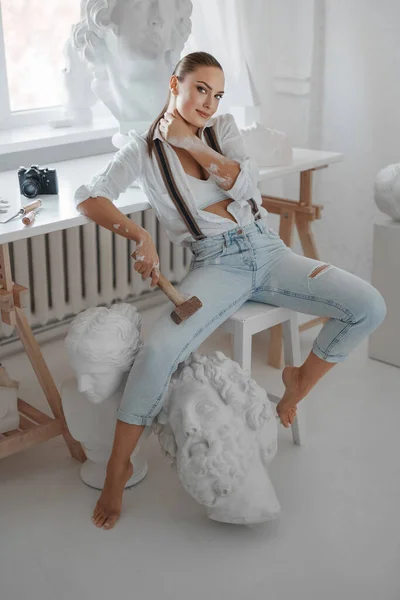 白いボタンなしのシャツとジーンズをサスペンサーで着た才能ある女性彫刻家は ハンマーを持ち 彫刻に囲まれた彼女の彫刻工房で椅子にポーズをかける — ストック写真