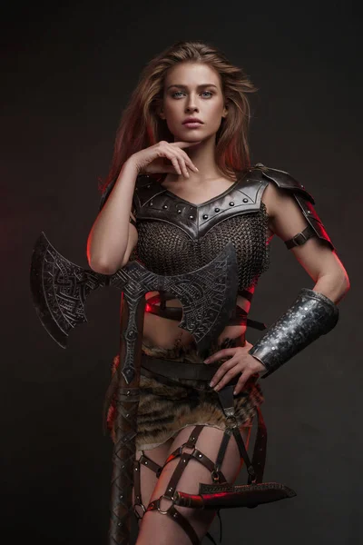 Stunning Viking girl dressed in a chainmail top and fur skirt poses with a two-handed axe against a textured gray wall