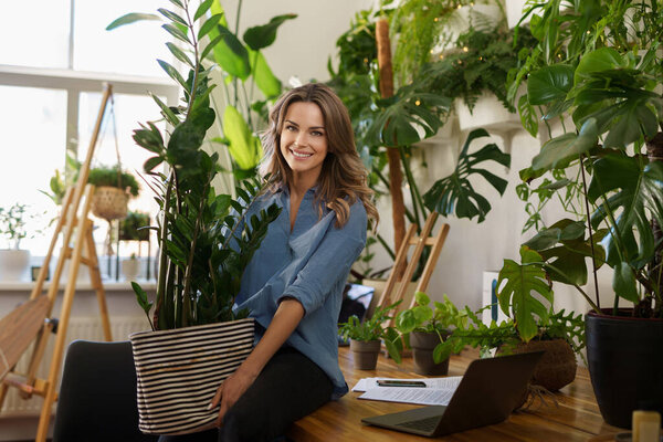 A smiling florist in a blue shirt holds a large plant pot in a room filled with plants and warm sunlight 
