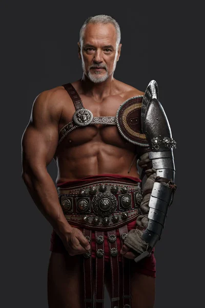 An elderly, yet muscular, gladiator is captured in historical armor set against a plain grey background