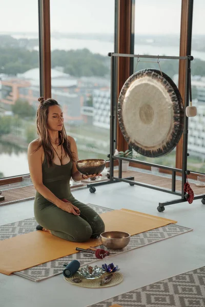 A serene atmosphere as a woman engages in spiritual practices, meditating with a singing Tibetan bowl in a city apartment amidst a picturesque urban backdrop