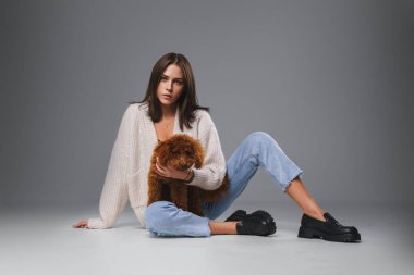 Göz kamaştırıcı bir esmer, yerde oturuyor, oyuncak kanişini sıkıca tutuyor, serin gri bir stüdyo tarafından çerçevelendiriliyor.