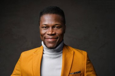 Yakışıklı, koyu tenli, parlak sarı ceketli, koyu dokulu bir arka plana gülümseyen bir adamın yakın çekimi.